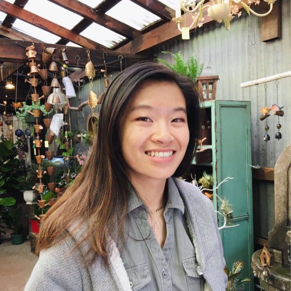 Image description: A woman smiling in a gray jacket in front of plants and hanging decorations.