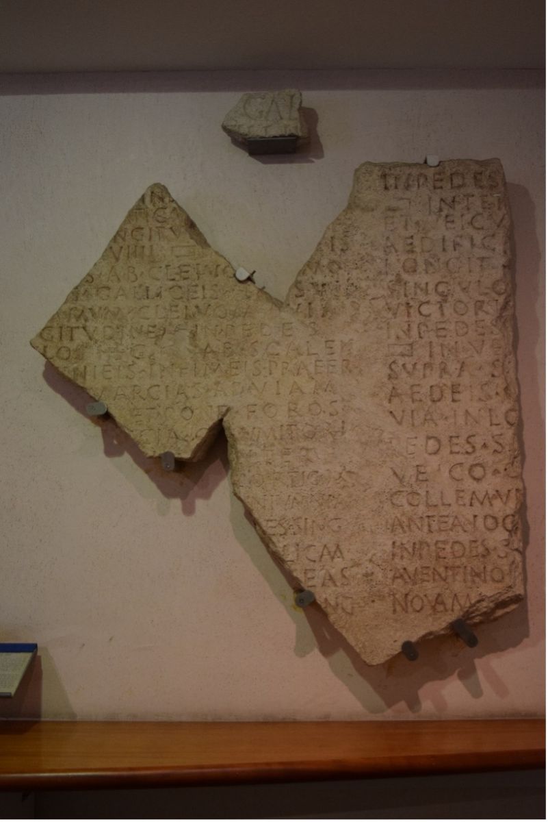 A beige tablet bearing a Latin inscription hangs on the wall of a museum. It is broken and has sections missing.