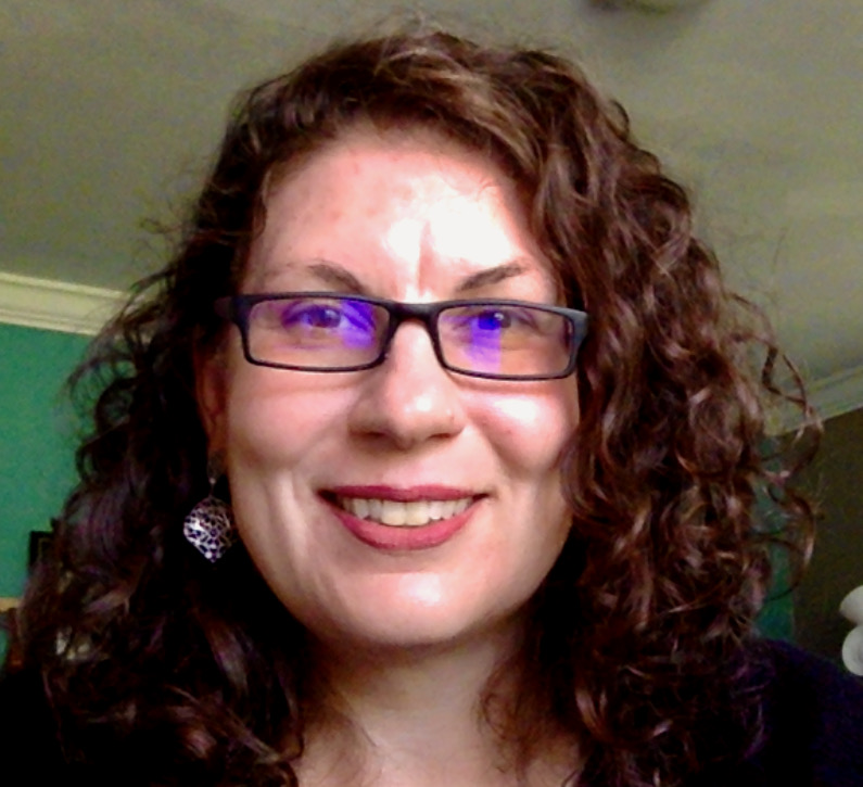 A white woman with shoulder-length, curly auburn hair and rectangular black glasses smiles at the camera.