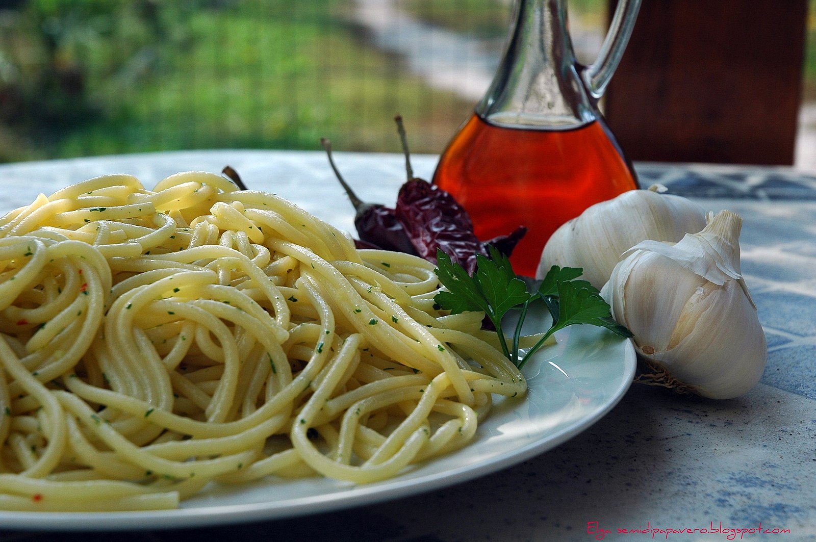 A photo of a plate of spaghetti with small green herbs scattered atop it. On the right side of the plate are a parsley spring and dried peppers. Next to the plate are two garlic bulbs and a decanter of vinegar.