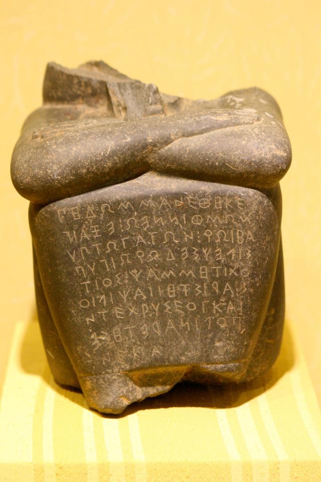 A dark stone statue with an Ionic Greek inscription on the front. Atop the inscription rest someone's folded arms. The top of the statue is broken off.