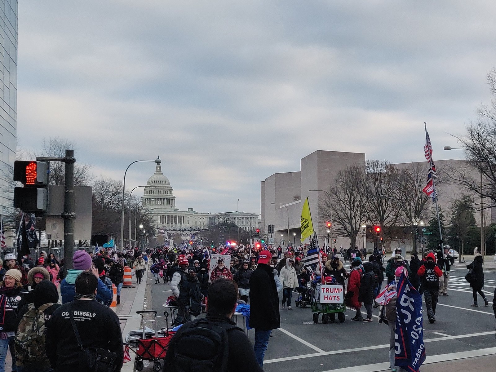 A crowd of people in the street leading to the Capitol building. Some carry flags and wear apparel with Trump's name and slogans.