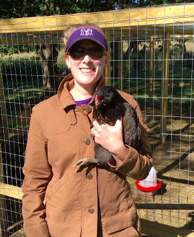 A white woman in a brown coat and purple visor holds a black bird in one hand and smiles at the camera.