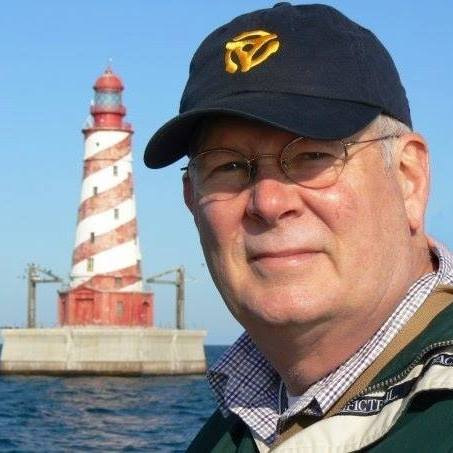 A light-skinned man wearing wire-frame glasses and a navy baseball cap looks at the camera. Behind him is a red and white lighthouse.