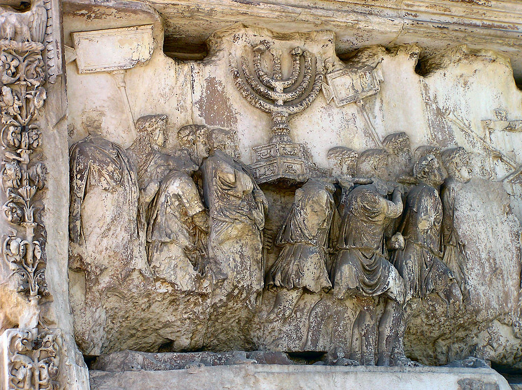 A stone carving illustrating a procession of men in drapery moving to the left, some of whom support the ark of the covenant, a large box atop which sits a menorah-like form.