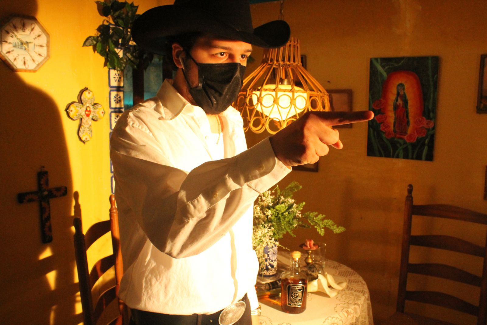 A man wearing a white dress shirt, a black cowboy hat, and a black face mask stands in a room with decorated walls. He points ahead of him.